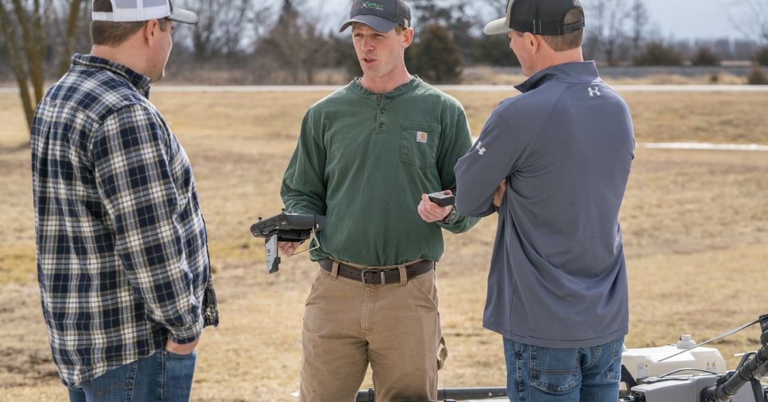 Three men standing in an open field next to an agricultural spray drone. The man in the middle holds the drone's controller.